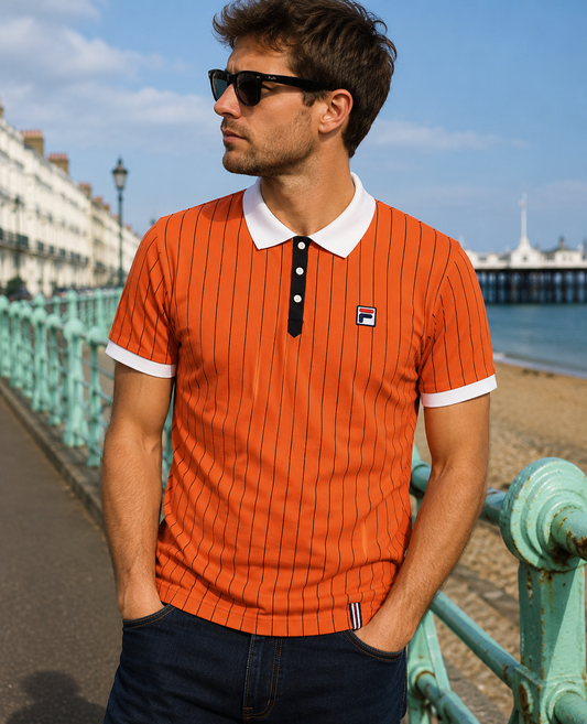 A man wearing an orange short-sleeve polo shirt with thin black pinstripes, a white collar, and a white button placket, standing on a seaside promenade. The shirt features a small Fila logo on the left chest.