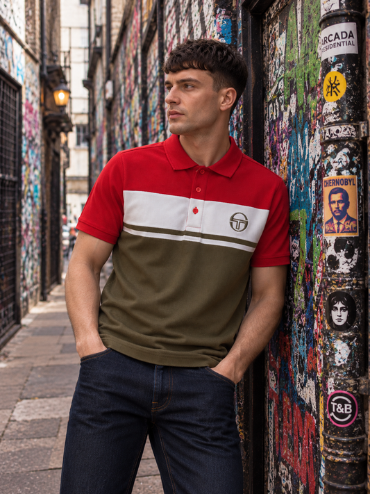 A man wearing a color-blocked polo shirt with red, white, and olive green sections, leaning against a graffiti-covered wall in an urban alley.