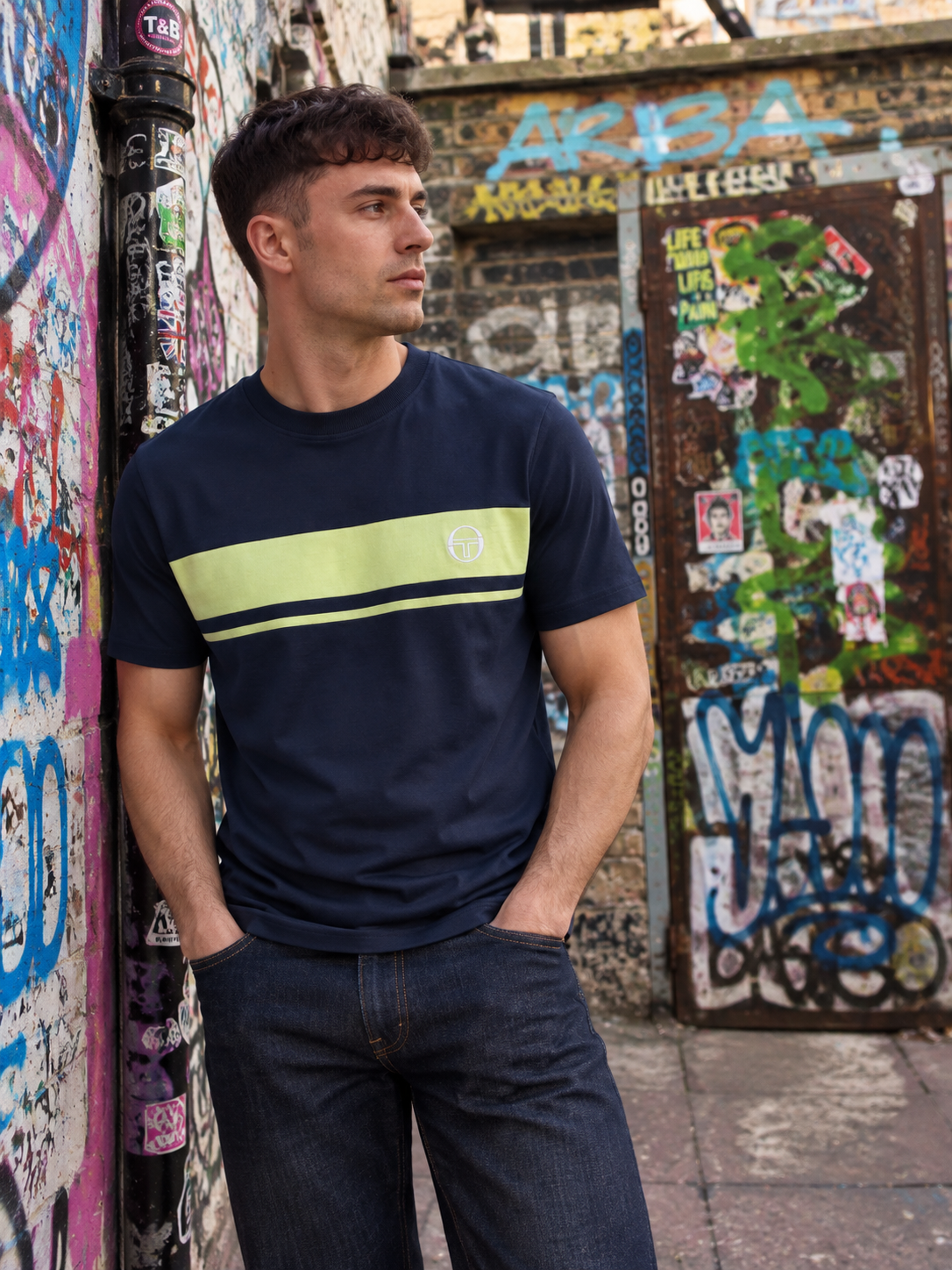 A man wearing a navy blue t-shirt with a lime green and black horizontal stripe across the chest, standing in an urban alleyway with graffiti-covered walls.