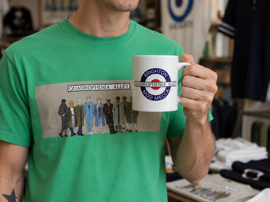 A person wearing a bright green graphic T-shirt holds up a white ceramic mug with a blue and red Brighton Wood Mecca logo. The background shows a blurred retail shop interior.