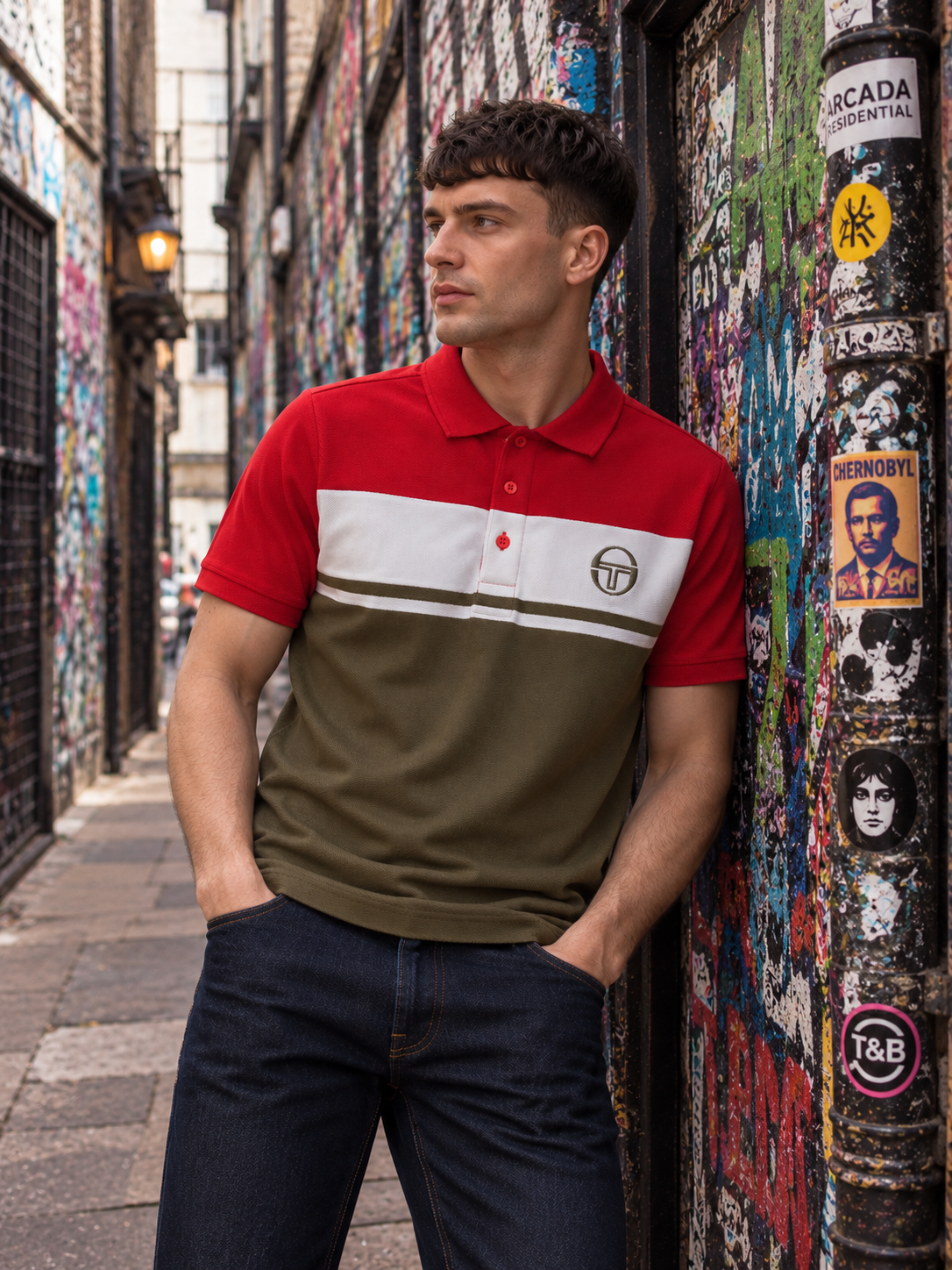 A man wearing a color-blocked polo shirt with red, white, and olive green sections, leaning against a graffiti-covered wall in an urban alley.