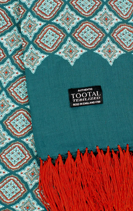 A close-up of a patterned scarf with teal, rust orange, and white geometric motifs, finished with a teal section and bright red fringe. A black woven label reads 'AUTHENTIC TOOTAL TEILIZED'.