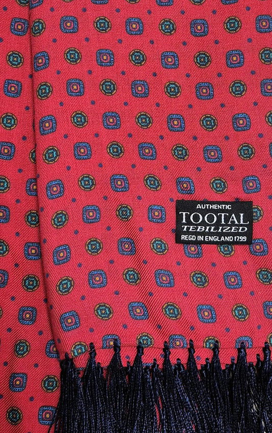 A close-up of a red woven scarf with a repeating geometric medallion pattern in blue, green, and yellow, finished with black fringe and a black woven label reading 'AUTHENTIC TOOTAL TEBILIZED REGD IN ENGLAND 1799'.